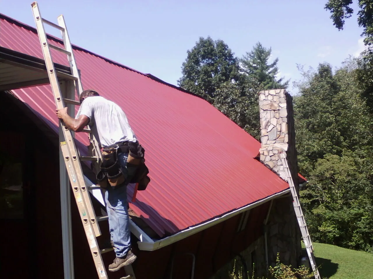 Roofer on a ladder beside a red metal roof and stone chimney — Soffit & Fascia Repair by Roofing Brothers in North Manchester